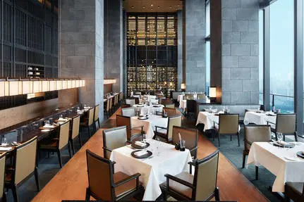 Aman Tokyo's dining venue with wooden flooring, white-dressed tables, and floor-to-ceiling windows overlooking the city.