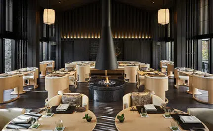 Aman Kyoto resort's Living Pavilion dining space with symmetrical wooden tables, central fireplace, and soft pendant lighting.