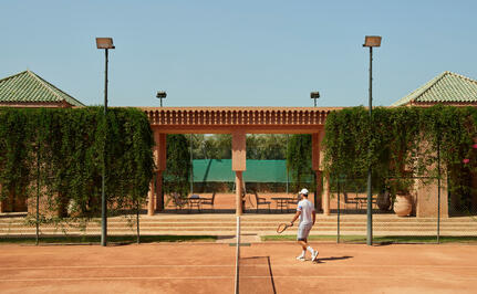 Amanjena clay tennis court with ivy-covered pavilion and verdant gardens under clear sky.