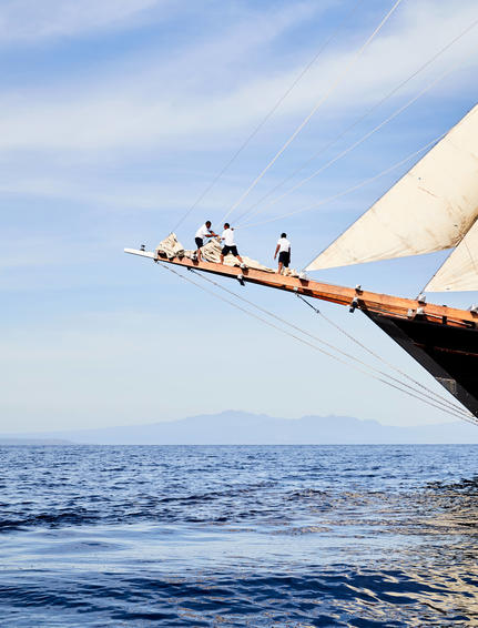 Amandira crew members perched on wooden boat bowsprit extending over calm blue sea.