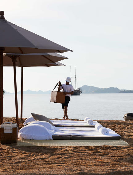 Woman carrying surfboard on beach at Amandira with calm waters and covered seating area in background.