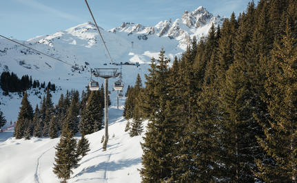 Ski lift ascending snowy slopes at Aman Le Mélézin, with snow-capped peaks and evergreen forest beyond.