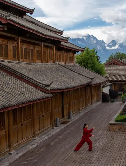 Monk in red robes walking across wooden deck at Amandayan, with traditional architecture and snow-capped mountains beyond.