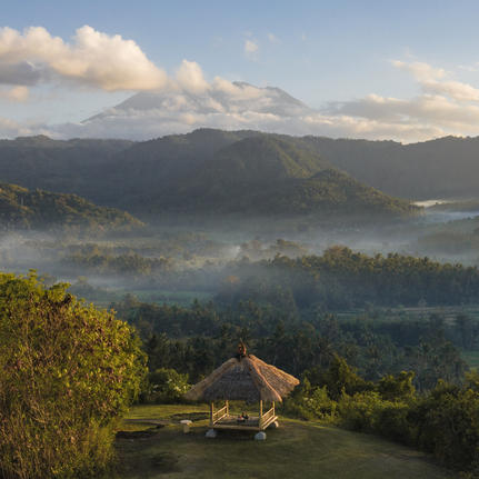 Amankila resort at sunrise, with mist rising over forested valleys and distant mountains under a pale sky.