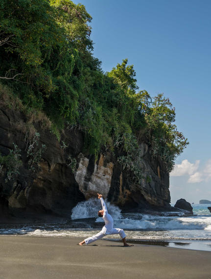 Woman in white standing beside traditional boat on sandy beach at Amankila, framed by lush cliff vegetation.