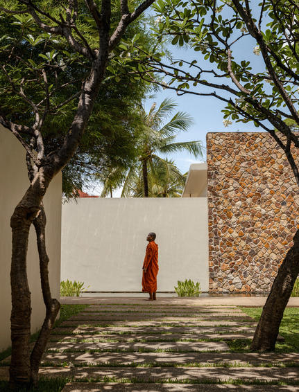 Monk in saffron robes walking through shaded courtyard at Amansara, Cambodia, framed by trees and stone walls.