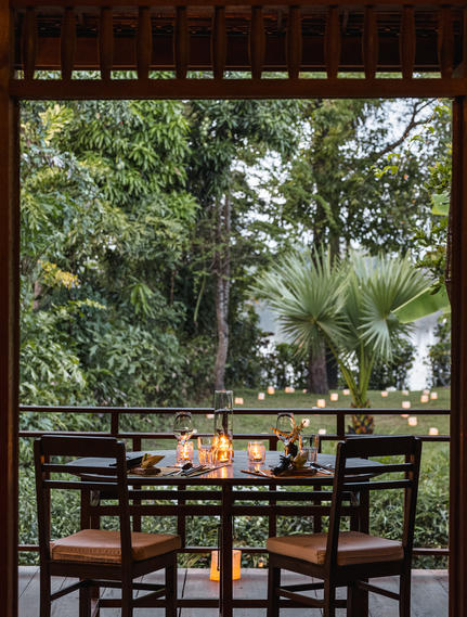Dining table set for two on a shaded terrace at Amansara, overlooking tropical gardens and palm trees.