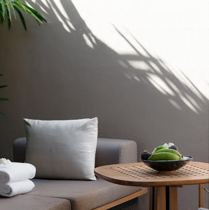 Amanwella suite detail showing cushioned seating, wooden side table with green vessel, and palm shadow on grey wall.