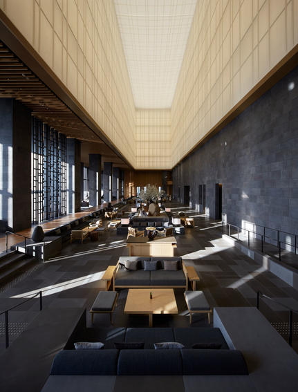 Aman Tokyo's lobby features a serene linear space with warm timber ceiling, dark stone walls, and carefully placed seating areas bathed in natural light.