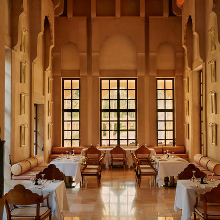 Amanbagh restaurant interior with warm stone walls, tall arched windows, and wooden dining chairs arranged throughout the space.