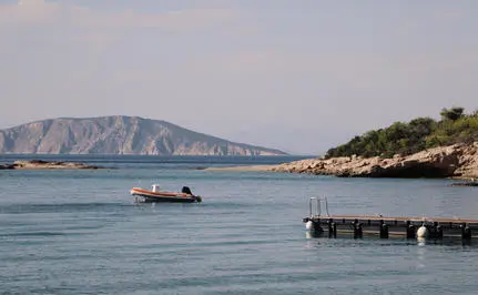 Wooden pier extending into clear turquoise waters at Amanzoe, Greece, with a small boat and distant cliffside visible.