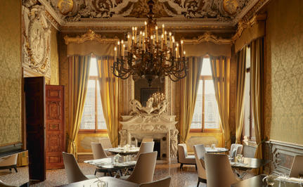 Ornate dining room at Aman Venice with gilded walls, crystal chandelier and tall windows draped in cream curtains.