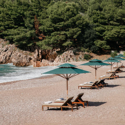 Sunlit beach at Aman Sveti Stefan with loungers beneath parasol, turquoise waters and rocky cliffs beyond.