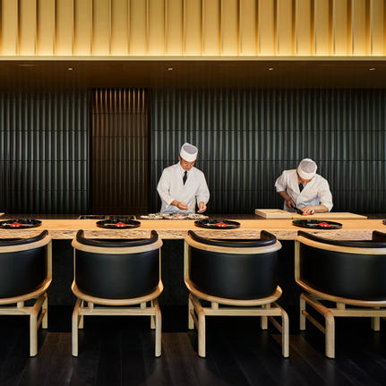 Two chefs stand behind a wooden counter at Aman Kyoto, with empty dining chairs lined in front.