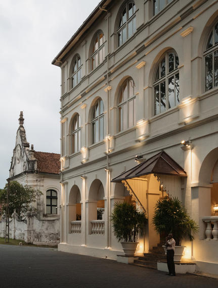 Amangalla's elegant façade at dusk, showcasing colonial architecture with illuminated ground-floor arches and a church spire beyond.