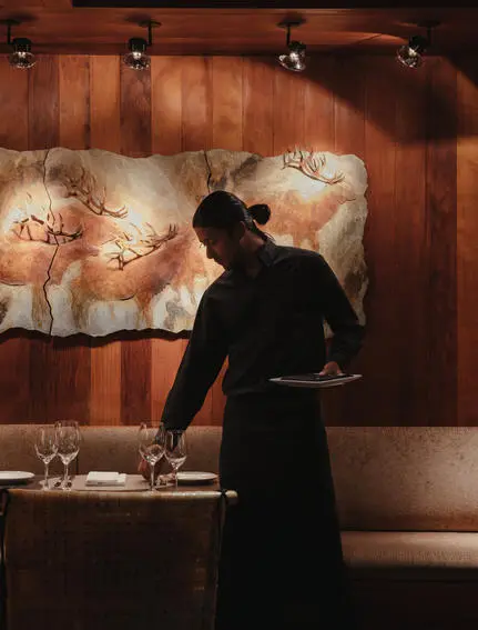 Server in black uniform arranging table settings in Amangani's dining venue, warm wood interior visible behind.