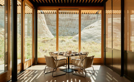 Camp Sarika restaurant at Amangiri, with floor-to-ceiling windows overlooking desert landscape, wooden table and chairs bathed in warm sunlight.