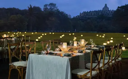 Tabel makan malam yang diterangi lilin di Amanjiwo resort, dengan pemandangan Candi Borobudur di latar belakang saat senja.