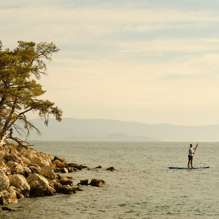 Person paddleboarding in calm turquoise waters at Amanruya, Turkey, with rocky shoreline and tree in foreground.