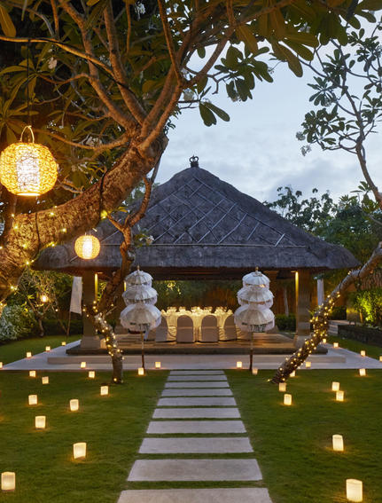 Candlelit pathway through manicured gardens at Aman Villas at Nusa Dua, leading to an illuminated pavilion at dusk.