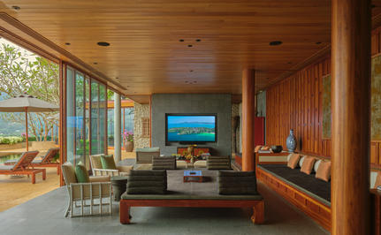 Amanpuri accommodation living room with warm wood panelling, floor-to-ceiling windows overlooking tropical landscape, and modern furnishings.