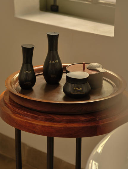 Aman Essentials skincare products displayed on a wooden round table at Amantaka, featuring black bottles and a cream jar.