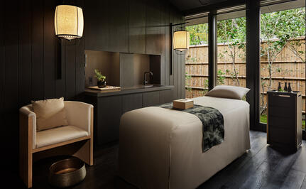 Serene wellness spa treatment room at Aman Kyoto with a massage bed, soft lighting, and garden views through large windows.