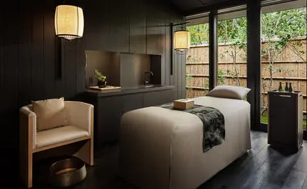 Salle de soins spa apaisante à Aman Kyoto avec lit de traitement, fauteuil crème et grandes baies vitrées donnant sur un jardin.