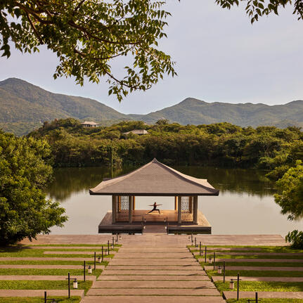 Wooden pavilion with pitched roof extending over still water at Amanoi, surrounded by verdant landscape and distant mountains.
