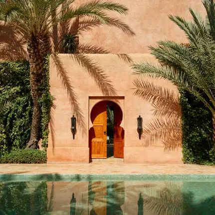 Architectural detail at Amanjena resort showing a terracotta archway with golden doors reflected in still water, flanked by palm trees.