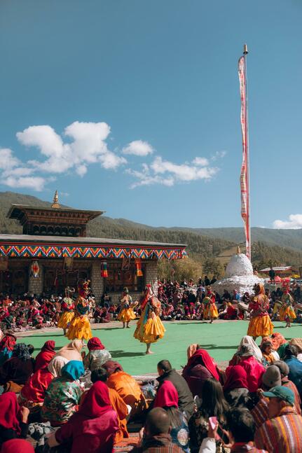 Colourful festival gathering at Amankora in Bumthang, with crowds seated on ground beneath clear blue sky.