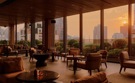 Aman Nai Lert Bangkok lounge with floor-to-ceiling windows overlooking Bangkok at sunset, warm lighting and contemporary furnishings.