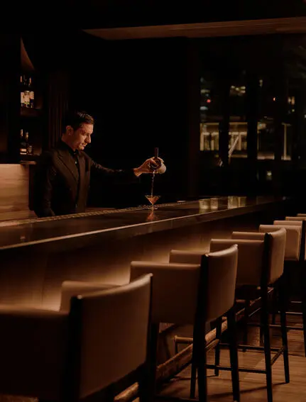 曼谷奈乐安缦酒店的酒廊，昏暗灯光下一名调酒师在吧台后方工作，木质吧凳整齐排列。