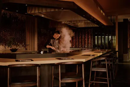 曼谷奈乐安缦酒店内，酒保在吧台后方调制鸡尾酒，昏黄灯光映照出温暖的氛围。