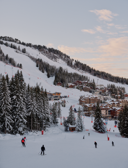 Skiers on snow-covered slopes at Aman Le Mélézin, with forested mountains and alpine buildings in the distance.