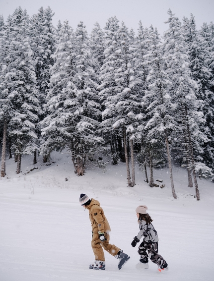 Skier and snowboarder walking through snow-covered pine forest at Aman Le Mélézin, France.
