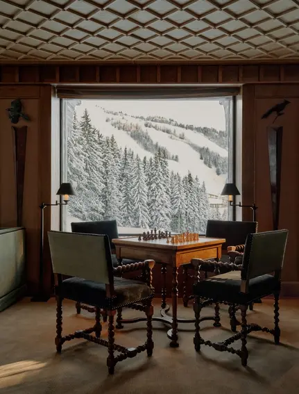 Salon at Aman Le Mélézin with wooden chairs at a desk facing a window overlooking snowy Alpine forest.