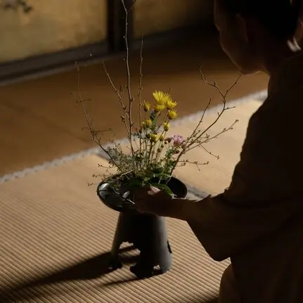 Arranging fresh flowers in a dark vessel at Aman Kyoto resort, with soft sunlight casting shadows across the tatami mat floor.