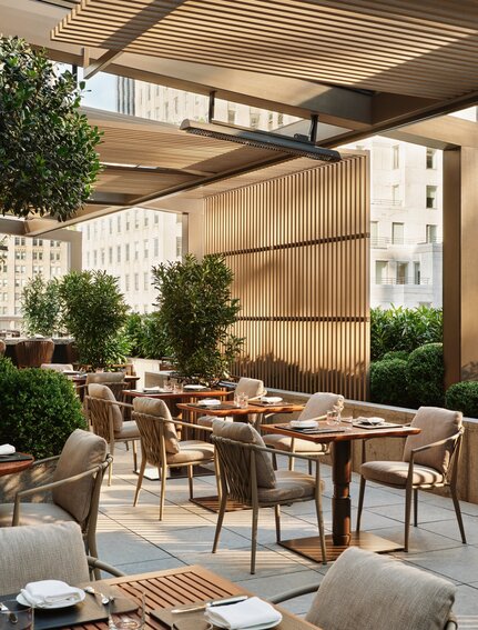 Terrazza del giardino di Aman New York con tavoli da pranzo in legno sotto una struttura coperta, circondata da piante e pareti in pietra.