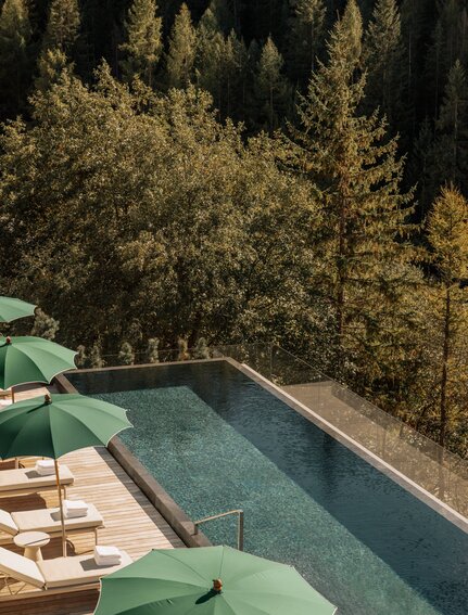 Outdoor swimming pool at Aman Rosa Alpina resort overlooking forested Italian Alps, with loungers and green umbrellas beside the water.