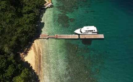 Wooden jetty extending into turquoise waters at Amanwana, Indonesia, with a white boat moored alongside, shot from above.