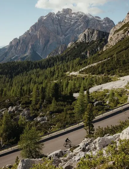 Cyclist on winding mountain road with peaks and forest at Aman Rosa Alpina resort.