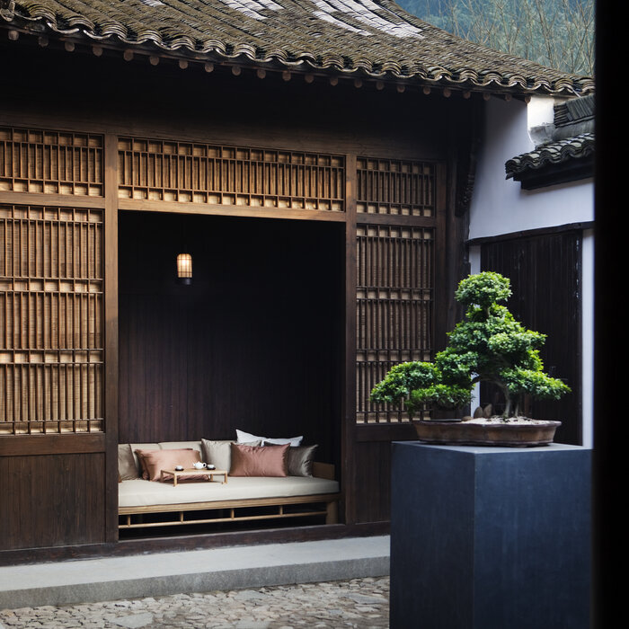 Amanfayun courtyard with dark wooden pavilion, white cushioned seating alcove, and potted bonsai tree.