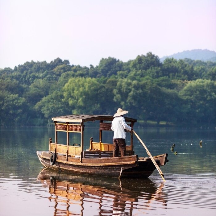 木质传统船只在西湖水面上，远处群山环绕，法云安缦度假村景色。