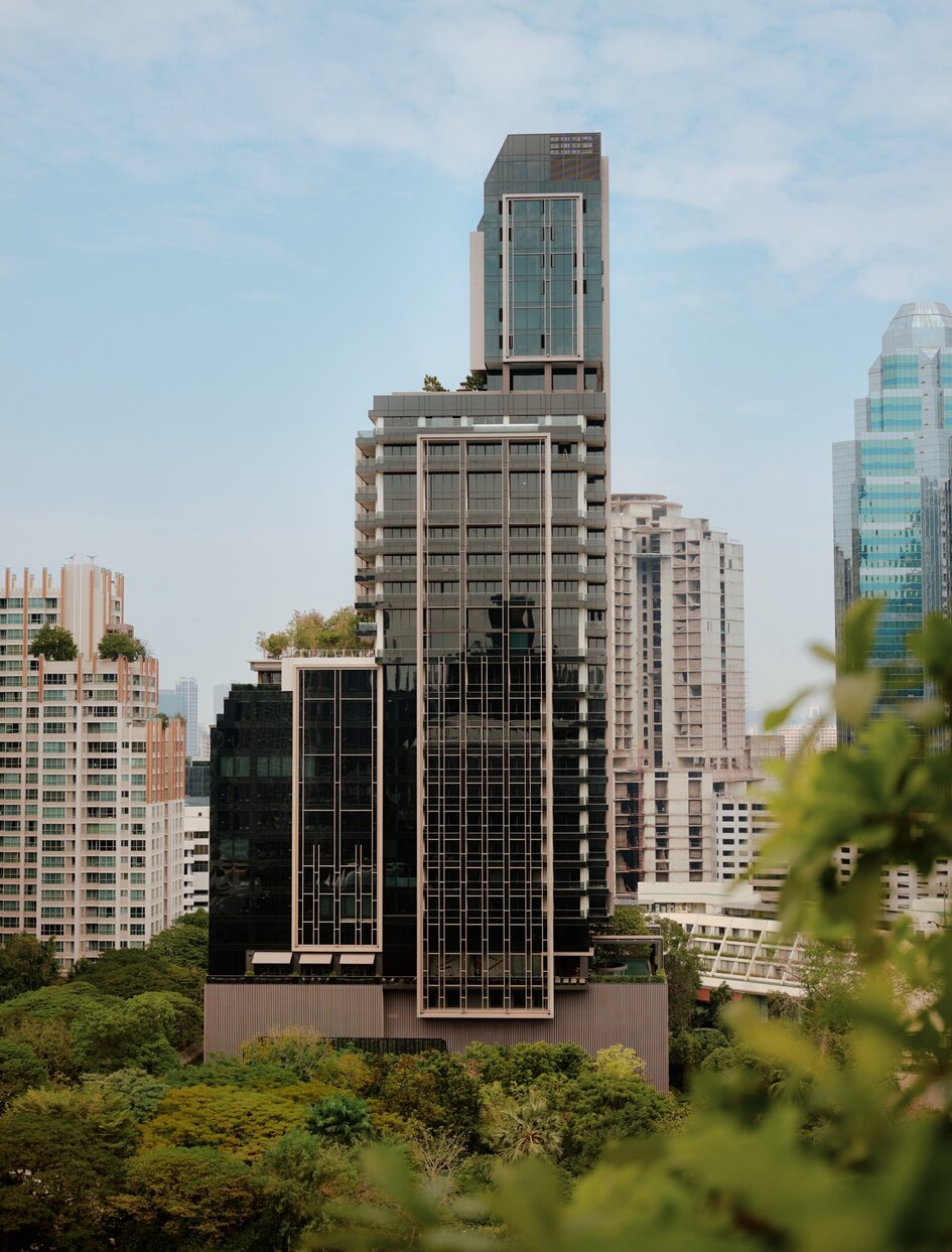 Aman Nai Lert Bangkok exterior with surrounding skyscrapers and green foliage in foreground.