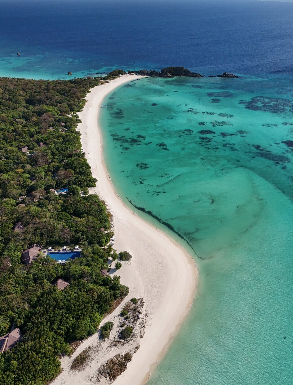 Aerial view of Amanpulo's crescent beach with turquoise waters, white sand, and forested shoreline.