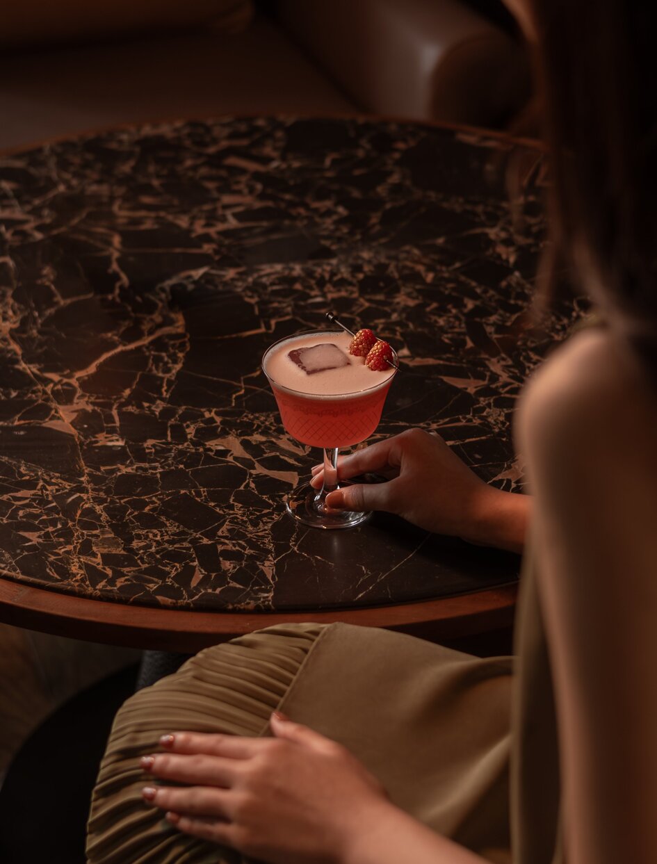 アマン ナイラート バンコクのラウンジで、コーラル色のカクテルを手に持つゲストの写真。