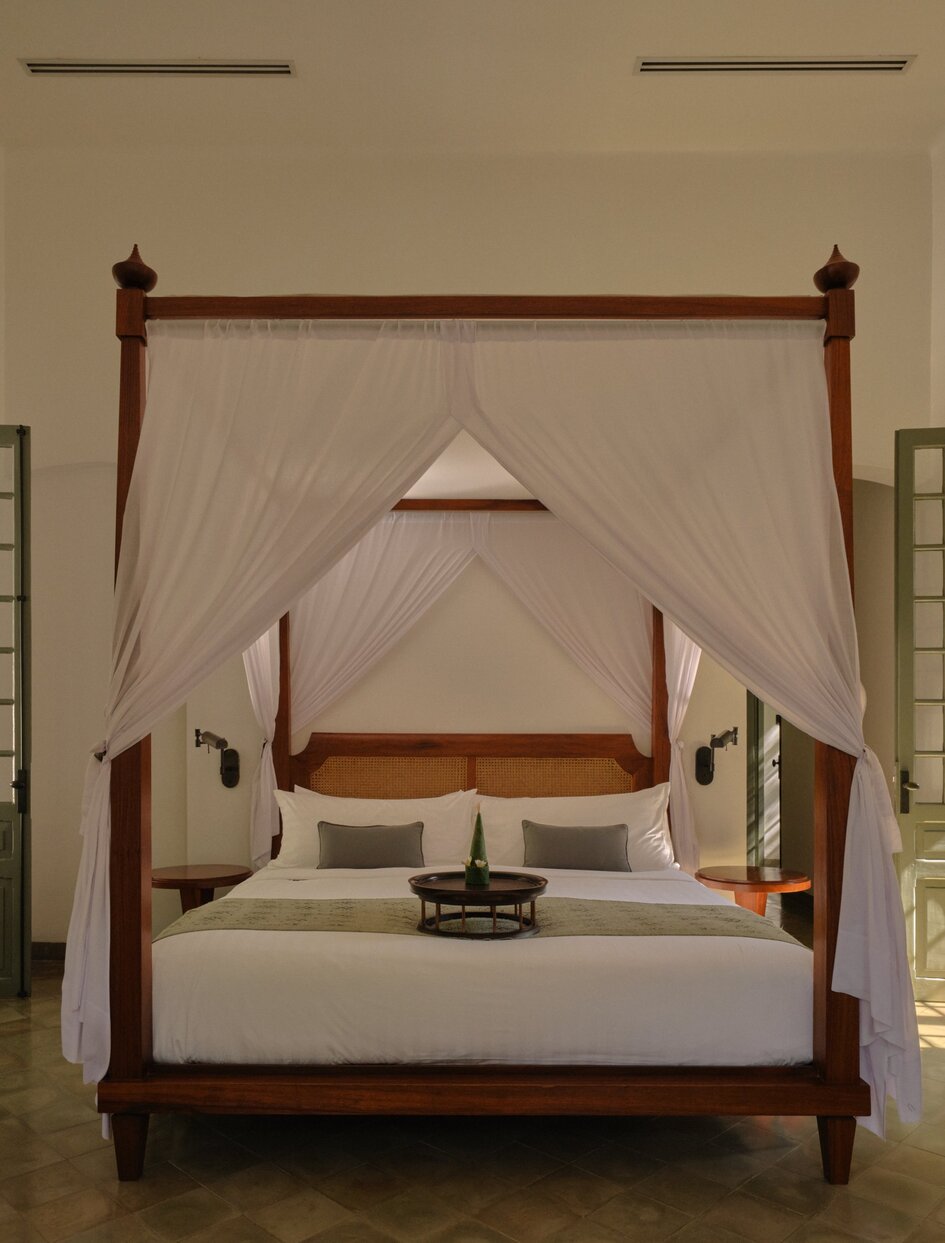 Amantaka bedroom with four-poster bed draped in sheer fabric, wooden furnishings and neutral tones.