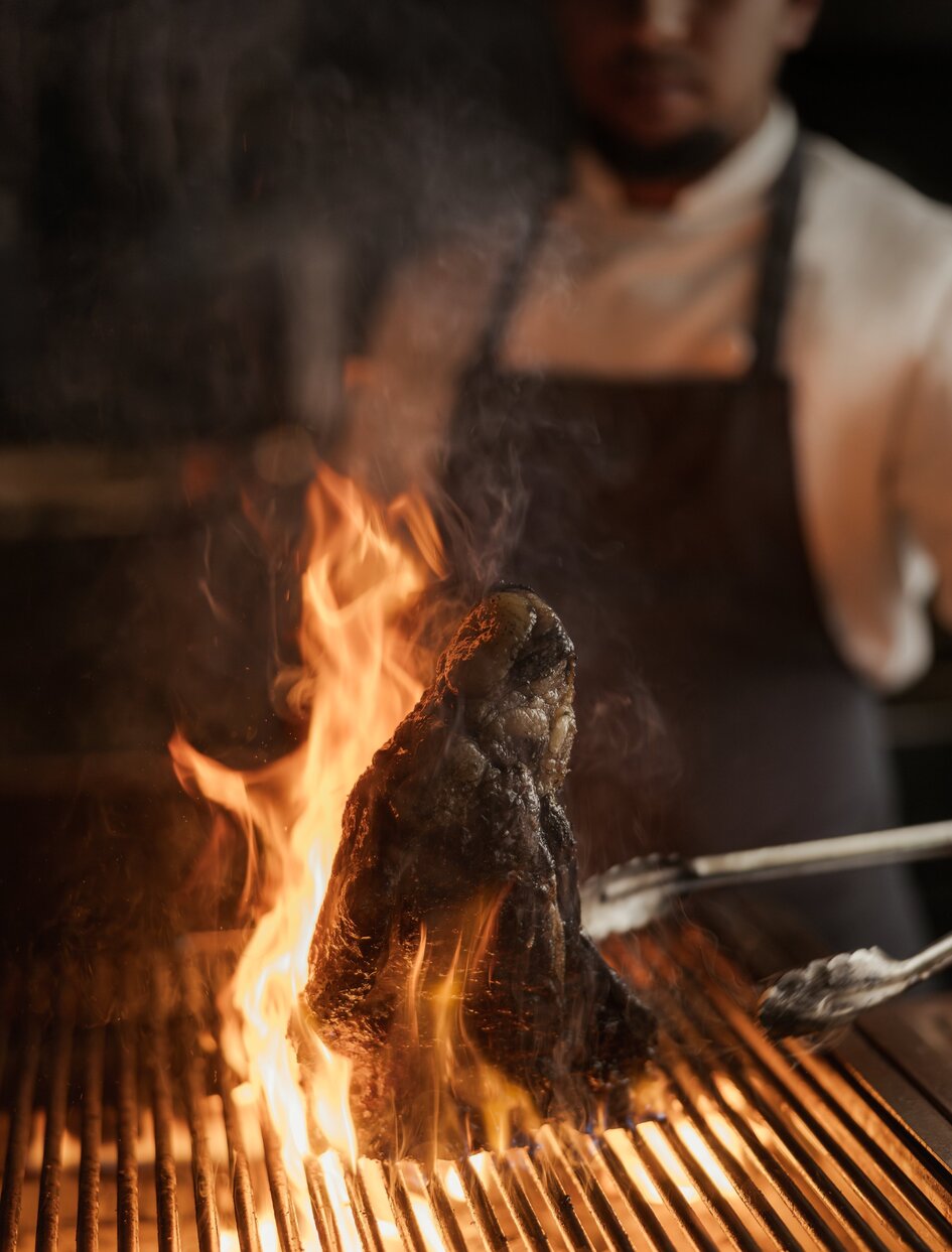 Chef grilling meat over open flame at Aman Rosa Alpina's grill restaurant in Italy.
