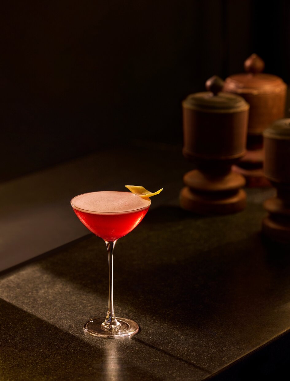 Amanoi bar cocktail in a coupe glass with red liquid, shaker and jigger in background.